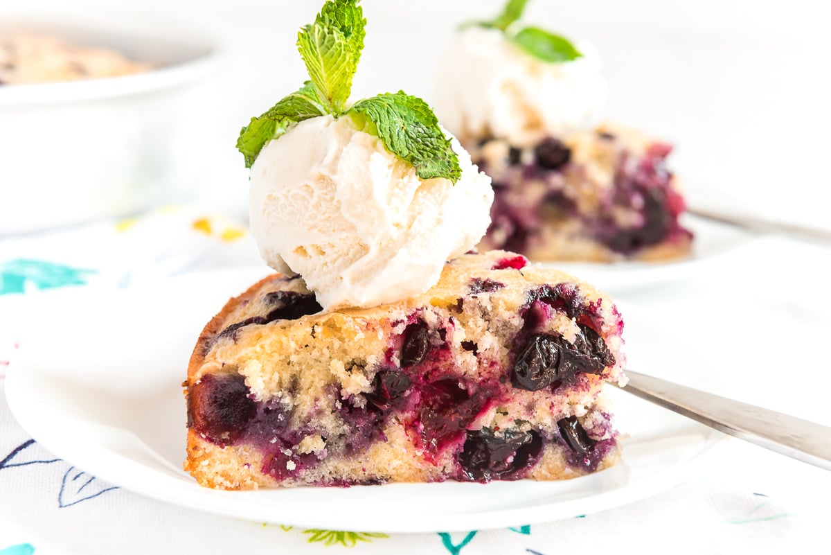 Slice of blueberry snack cake on a white plate topped with ice cream and garnished with mint.