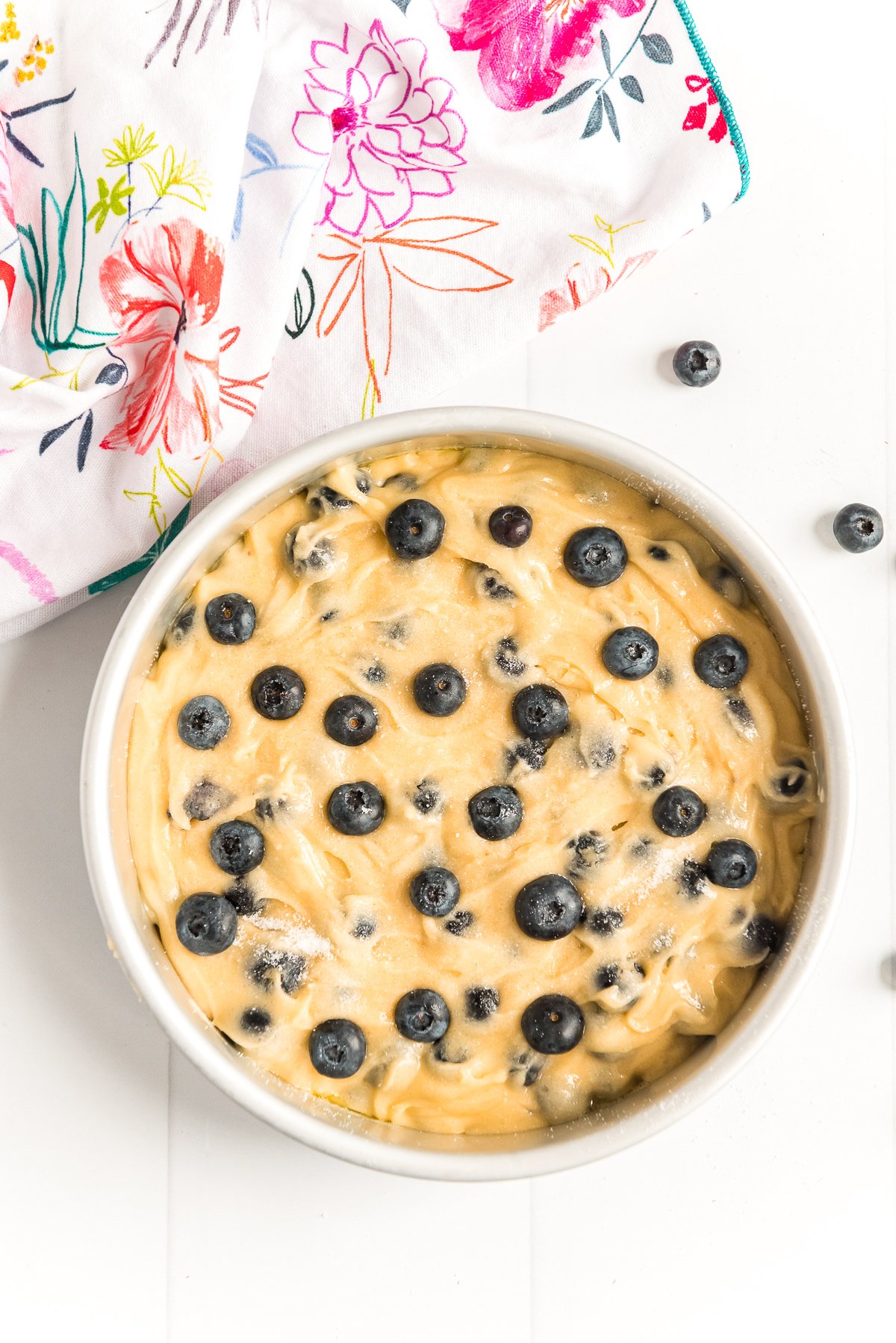 Blueberry cake batter in a round cake pan with a colorful floral napkin next to it.