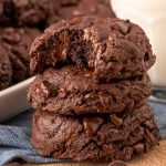 A stack of three double chocolate chip cookies on a blue napkin with the top one missing a bite.