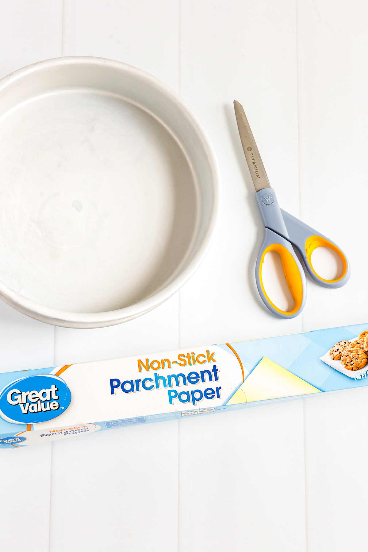 Cake pan, roll of parchment paper, and pair of scissors sitting on a white wooden table.