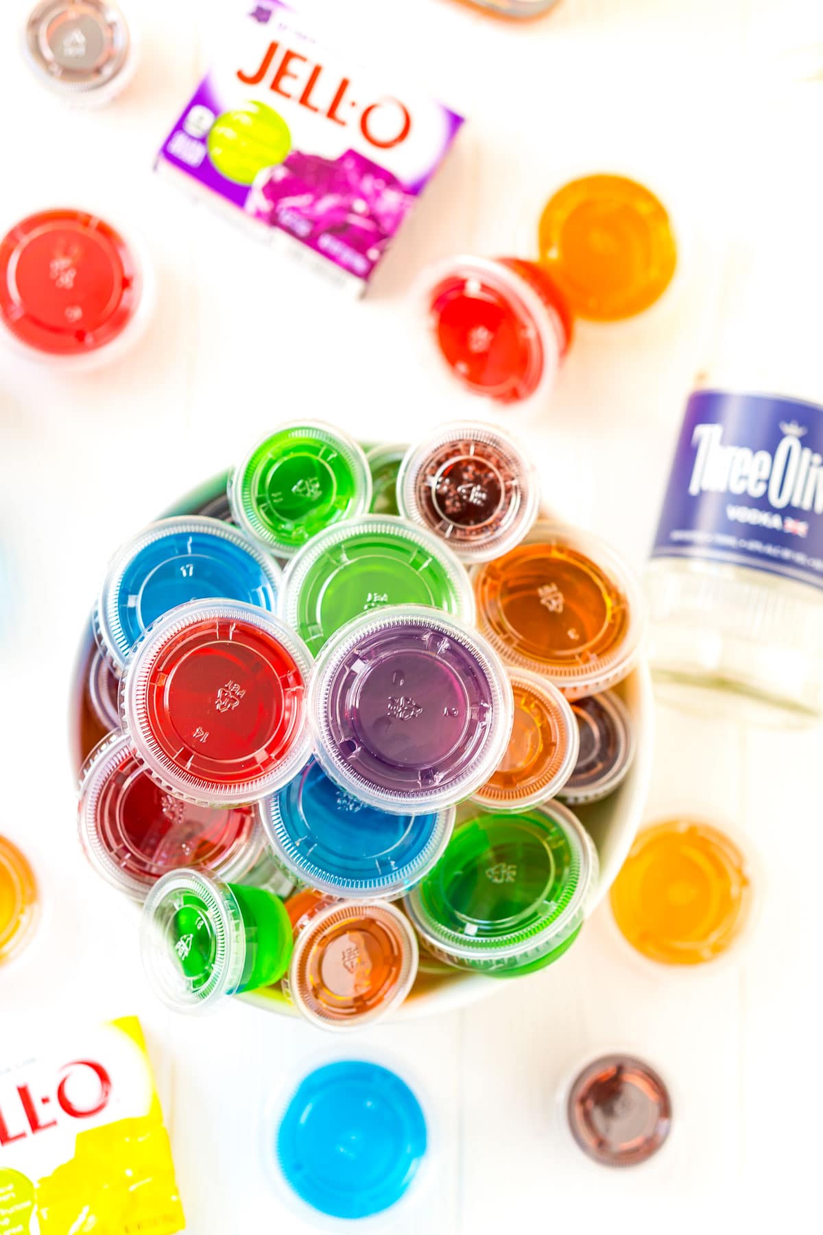 Bowl full of multi-colored jello shots with more jello shots, boxes of Jell-O, and a bottle of vodka around it.