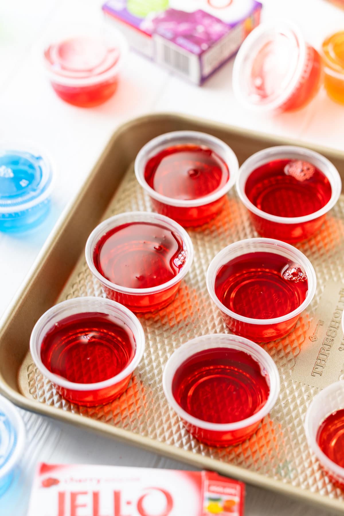 Red Jello Shots sitting on a baking sheet surrounded by boxes of jello and other jello shots.