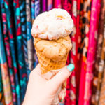 Woman holding up an ice cream cone in front of colorful scarves.