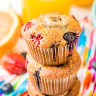 Three berry muffins stacked on top of each other on a colorful napkin with a glass of orange juice in the background.