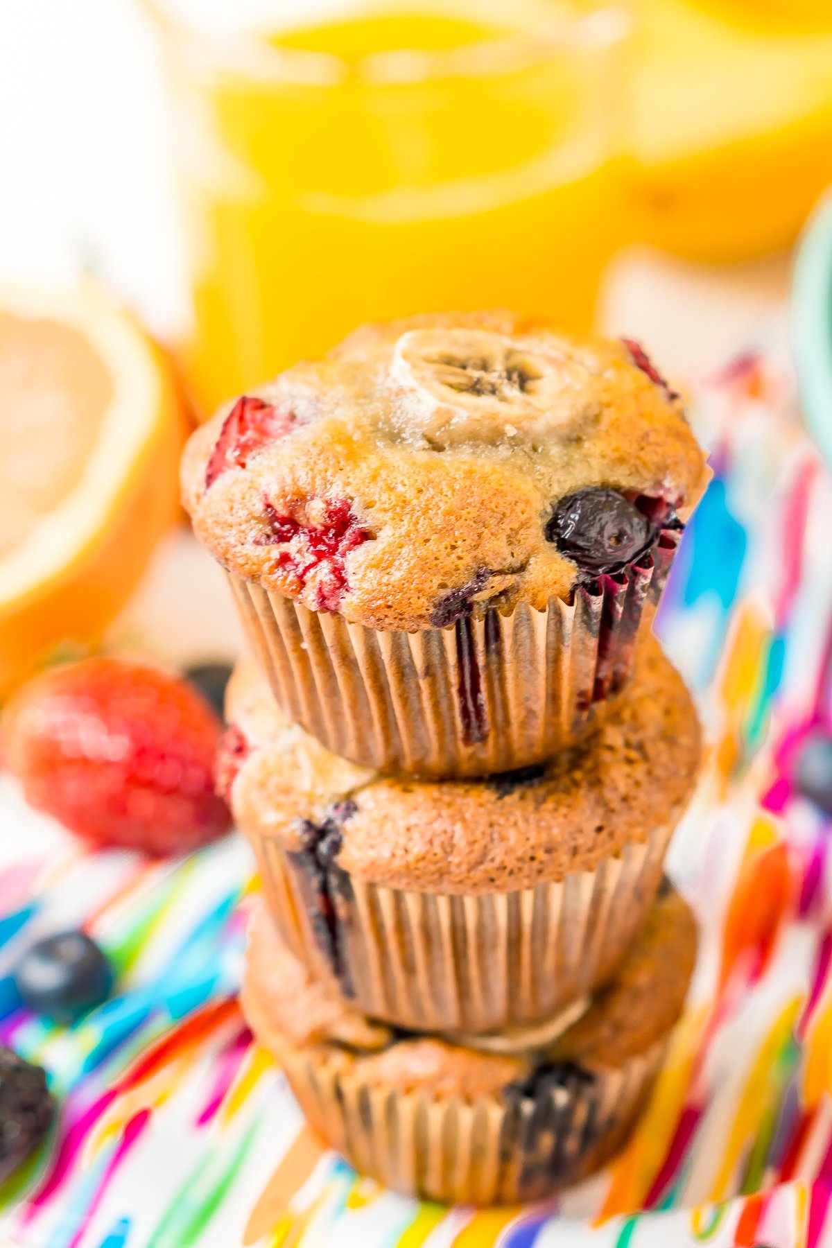 Three berry muffins stacked on top of each other on a colorful napkin with a glass of orange juice in the background.