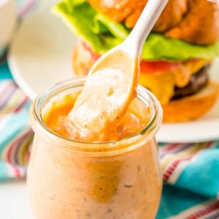 White spoon being pulled out of a small glass jar of burger sauce.