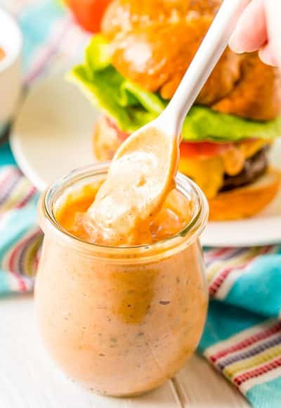 White spoon being pulled out of a small glass jar of burger sauce.