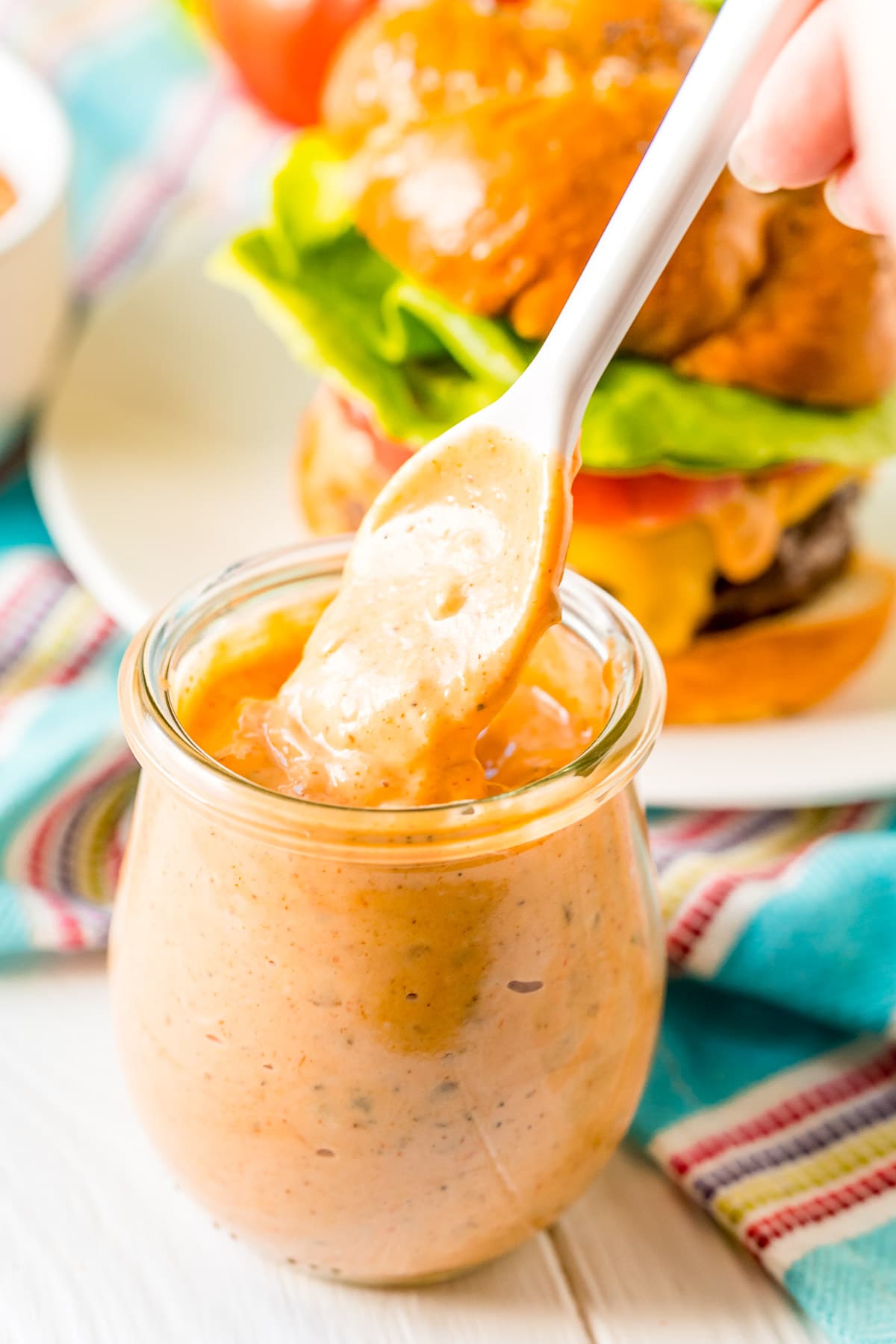 White spoon scooping burger sauce out of a small glass jar. Burger and blue striped napkin in the background.