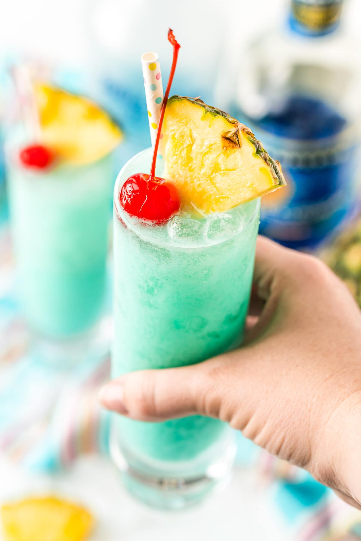 Woman's hand holding a blue hawaiian cocktail garnished with pineapple wedges and a maraschino cherry.