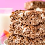 Stack of three cocoa pbbles no bake treats on a white plate on a colorful napkin with a glass of milk in the background.