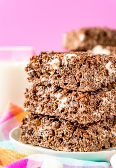 Stack of three cocoa pbbles no bake treats on a white plate on a colorful napkin with a glass of milk in the background.