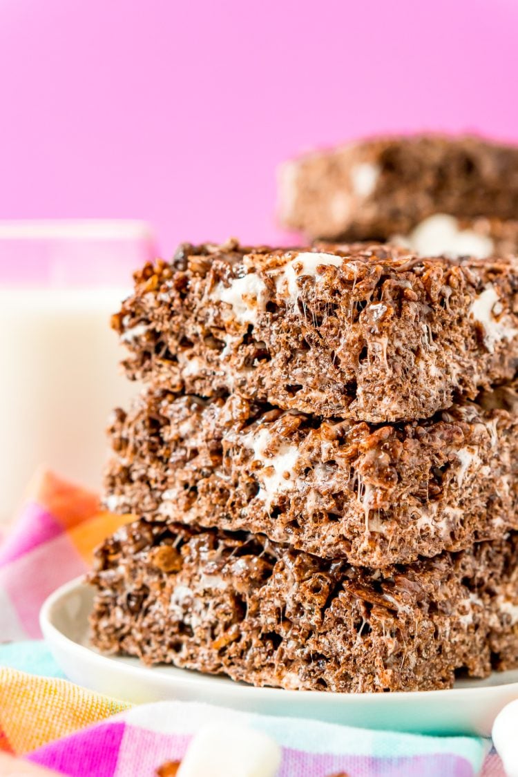 Stack of three cocoa pbbles no bake treats on a white plate on a colorful napkin with a glass of milk in the background.