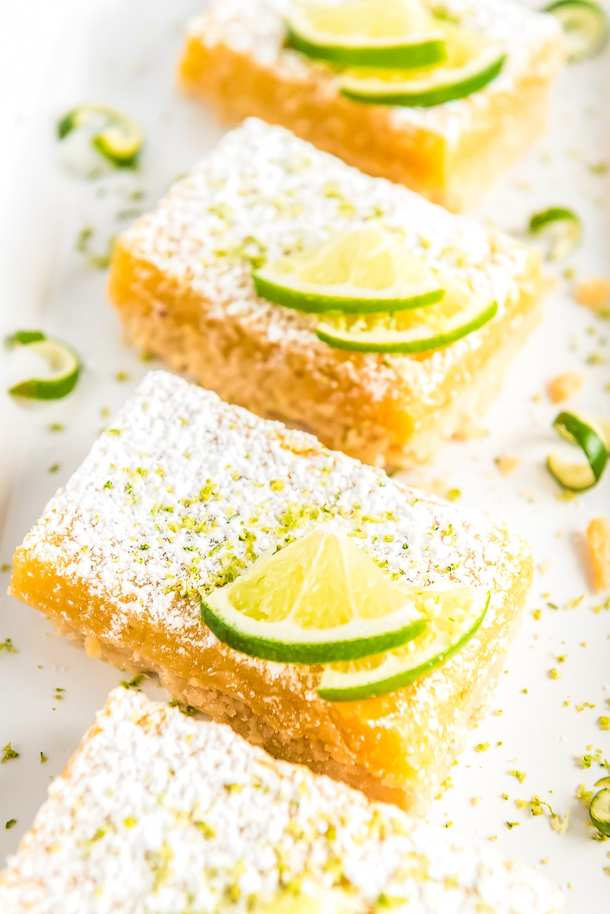 Lime Bars lined up on a white plate topped with lime wedges and powdered sugar.