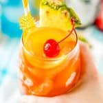 Woman's hand holding a glass filled with a rum runner cocktail garnished with orange, pineapple, and cherry.