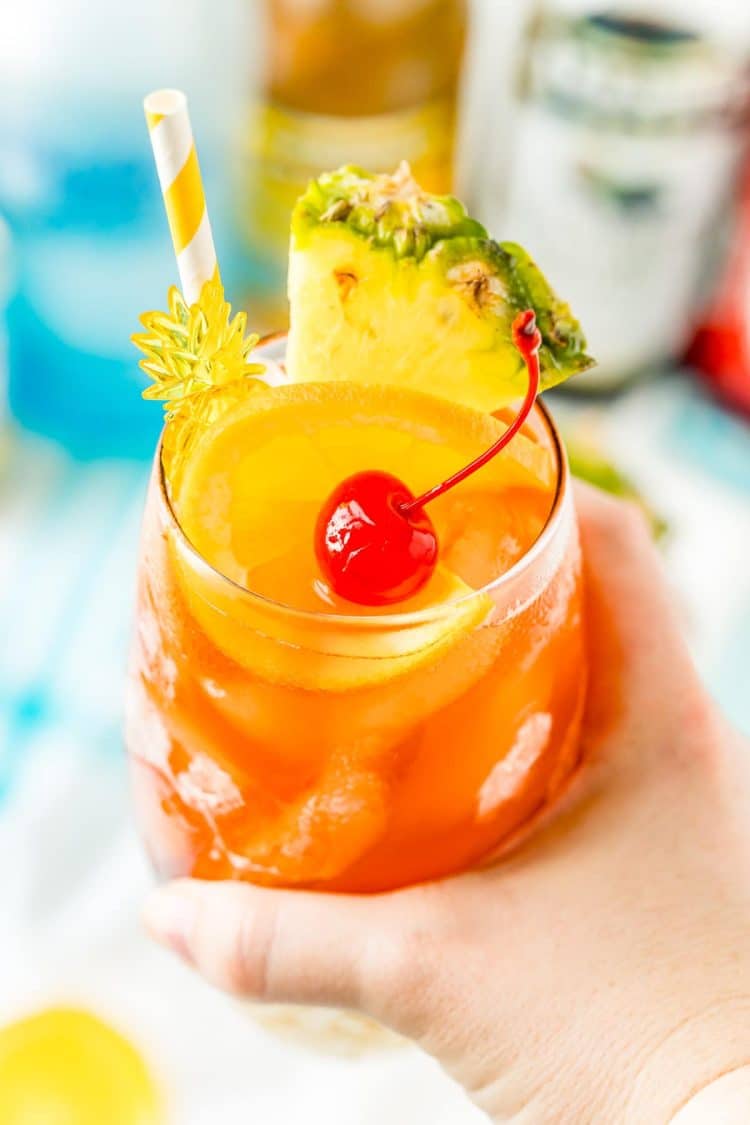 Woman's hand holding a glass filled with a rum runner cocktail garnished with orange, pineapple, and cherry.