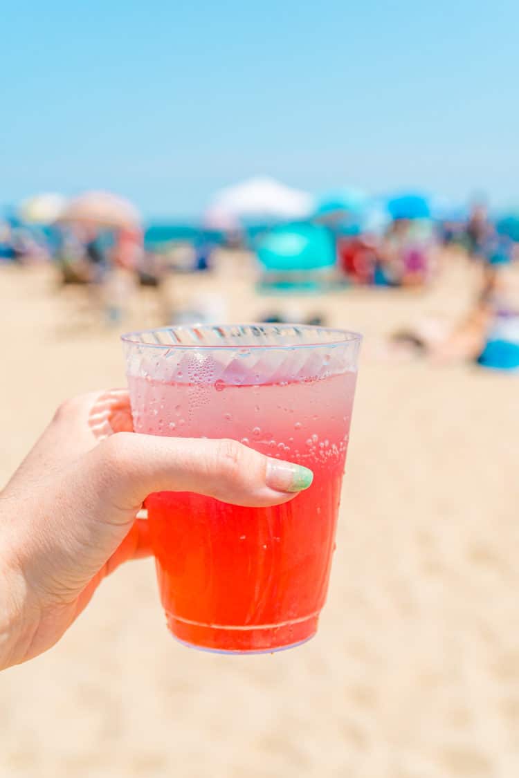 Woman's hand holding a cup of Cranberry Lemonade at the beach.