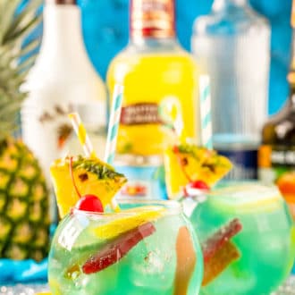 Fish bowl cocktail with pineapple and bottles of alcohol in the background.
