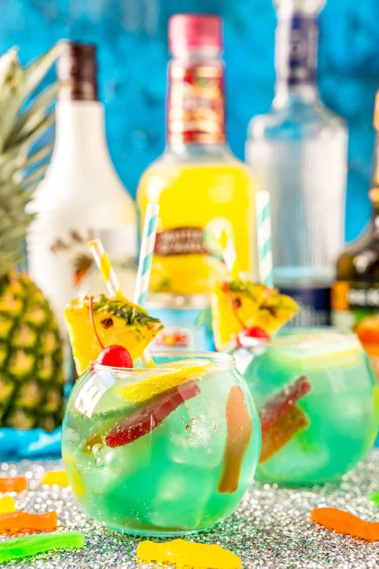 Fish bowl cocktail with pineapple and bottles of alcohol in the background.