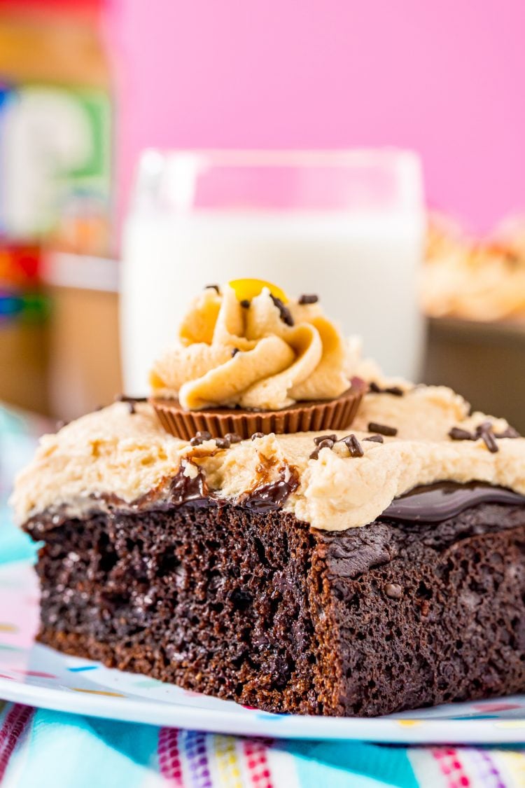 Close up photo of a slice of chocolate cake with peanut butter frosting on a white plate with a glass of milk in the background.