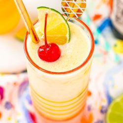 Close up photo of a pineapple daiquiri garnished with a maraschino cherry, lime wedge, and gold straw.