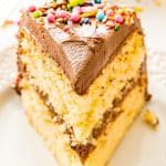 Slice of yellow cake with chocolate frosting and sprinkles on a white plate.