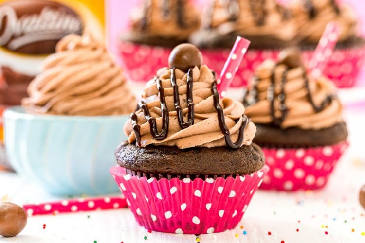 Photo of chocolate cupcakes and a small bowl of malt frosting behind them. Jar of Ovaltine on the counter in the background.