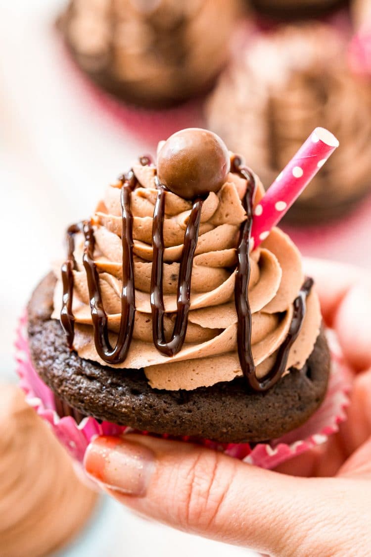 Woman's hand holding a chocolate cupcake drizzled in chocolate sauce.