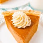 Close up photo of a slice of pumpkin pie on a white plate topped with whipped cream.