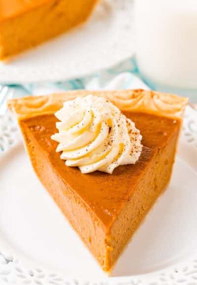 Close up photo of a slice of pumpkin pie on a white plate topped with whipped cream.