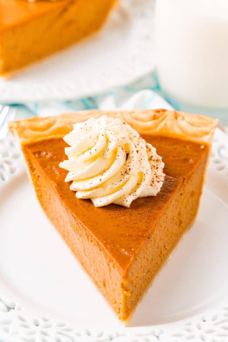 Close up photo of a slice of pumpkin pie on a white plate topped with whipped cream.