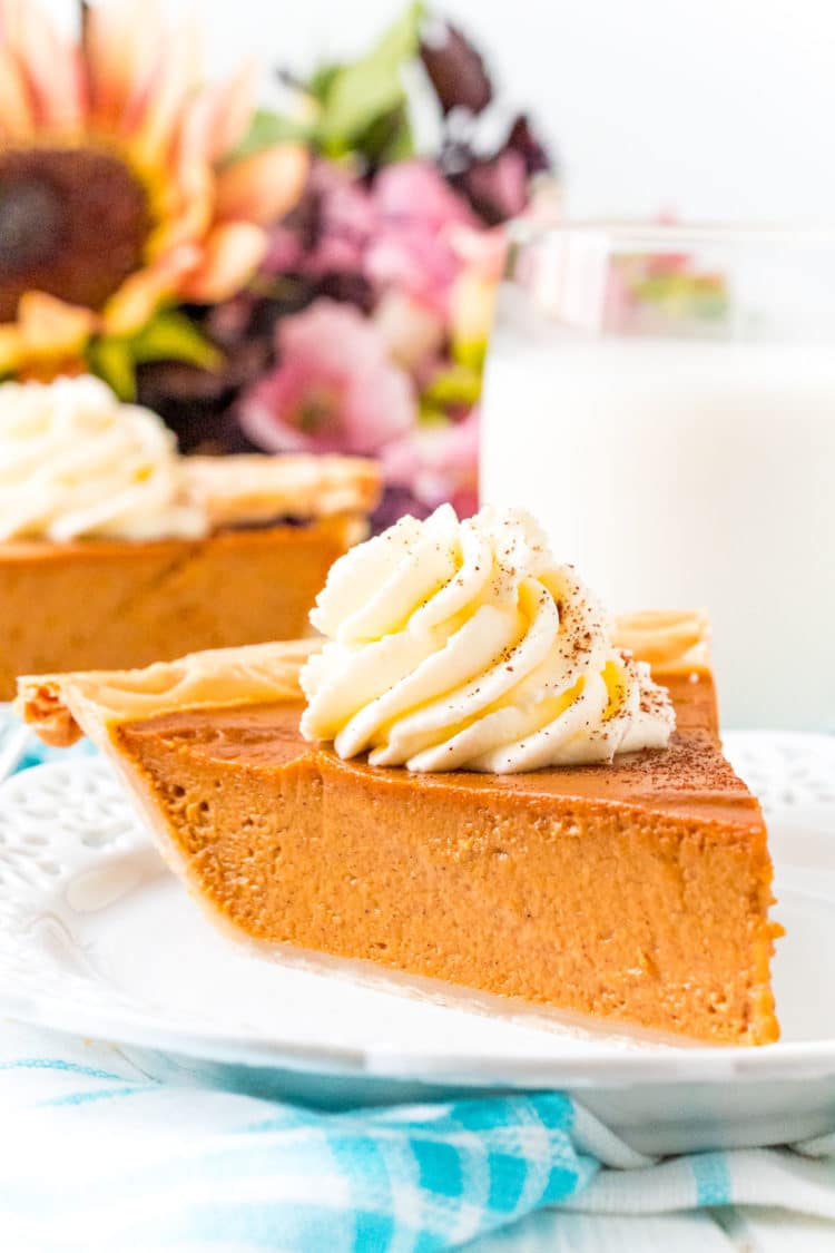 Photo of a slice of pumpkin pie on a white plate with a glass of milk and flowers in the background.