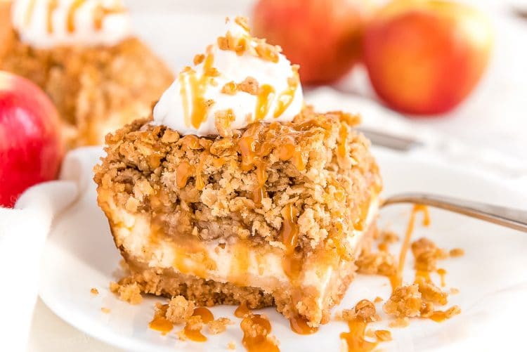 A square slice of an apple cheesecake bar on a white plate drizzled with caramel sauce. Apples and more slices of cheesecake are in the background.