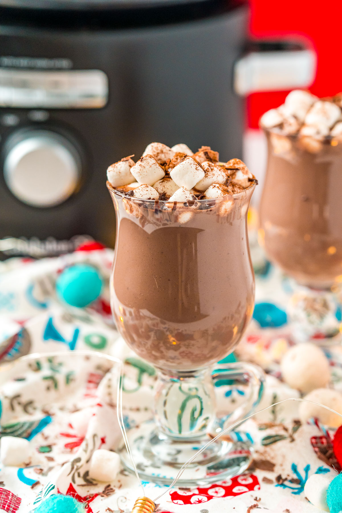 Two mugs of crockpot hot chocolate topped with marshmallows sitting on a festive napkin in front of a crockpot.