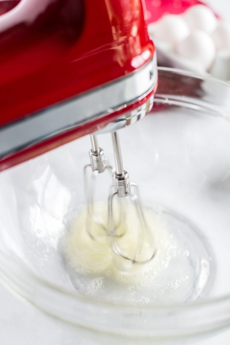 Egg whites being beaten with a hand mixer in a glass bowl.