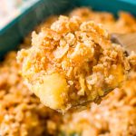 A wooden serving spoon lifting a scoop of pineapple casserole out of a serving dish.