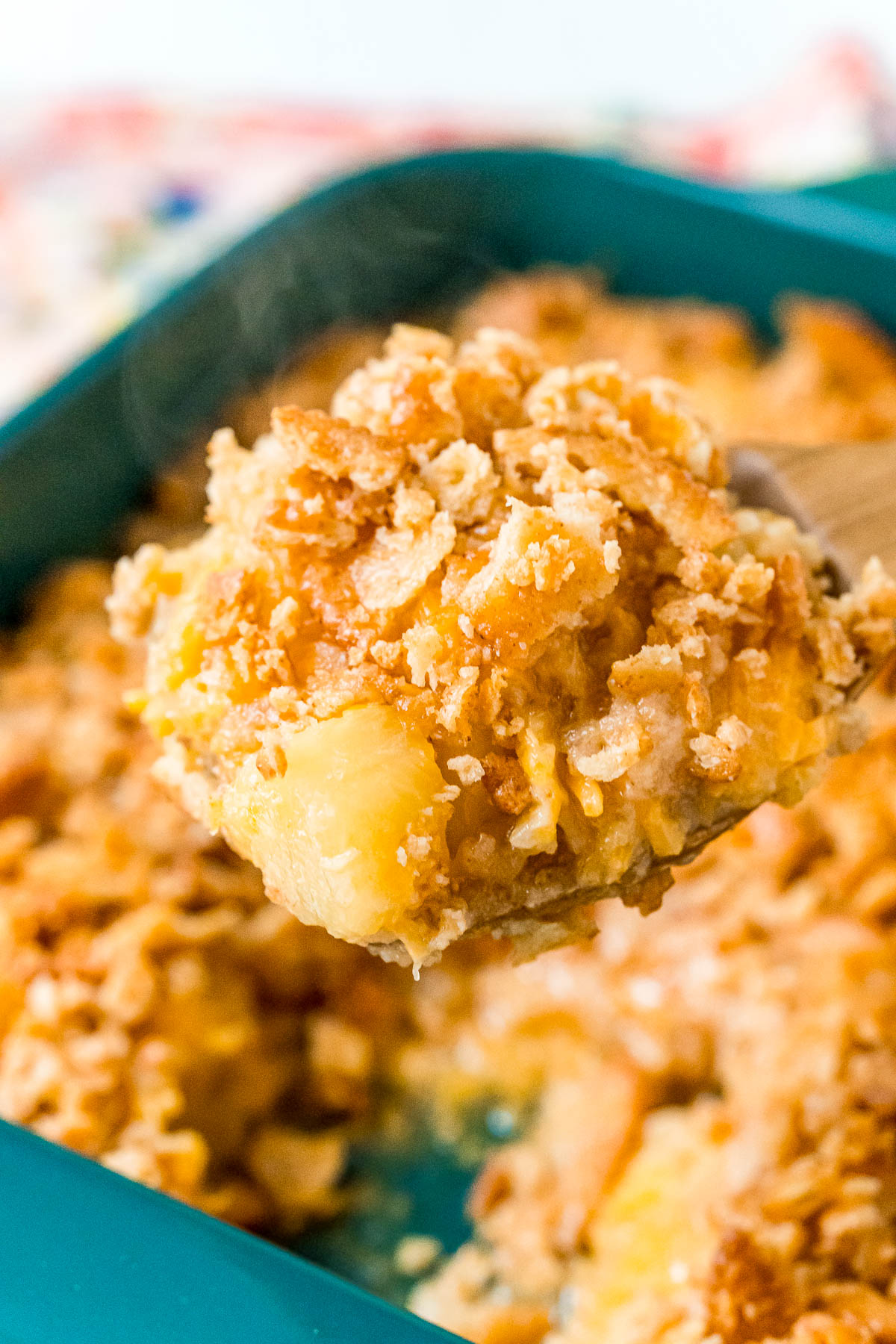 A wooden serving spoon lifting a scoop of pineapple casserole out of a serving dish.