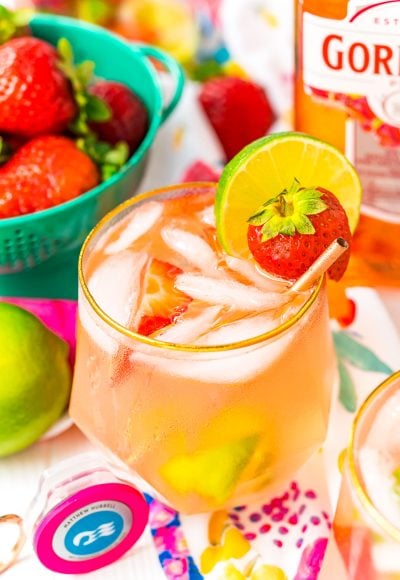 Close up photo of a pink gin spritz in a gold rimmed glass garnished with a lime and strawberry.