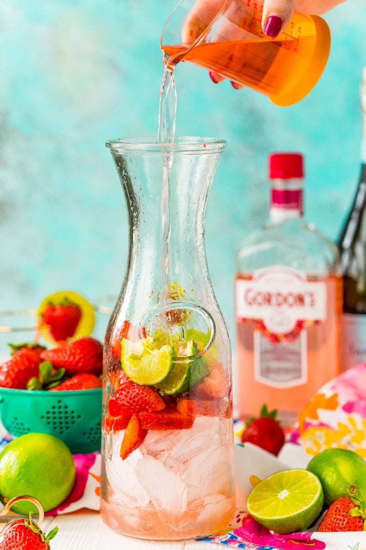 Pink liquor being poured into a carafe with strawberries and limes.