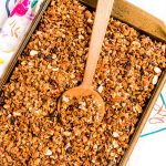 Homemade granola on a sheet pan with a wooden spoon scooping it.