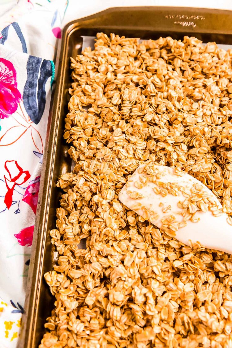 Oats being spread out on a large baking sheet with a rubber spatula to make granola.