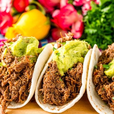 Close up photo of three barbacoa tacos on a cutting board with pink flowers in the background.