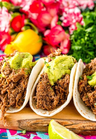 Close up photo of three barbacoa tacos on a cutting board with pink flowers in the background.