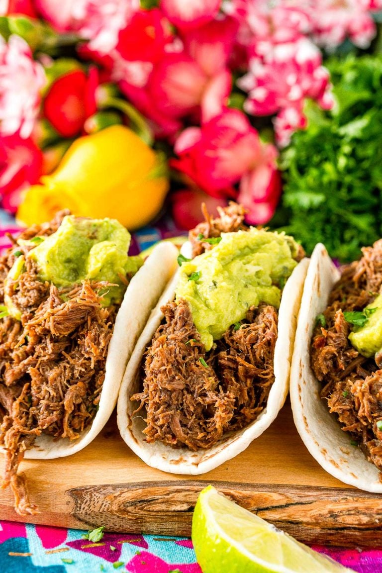 Close up photo of three barbacoa tacos on a cutting board with pink flowers in the background.