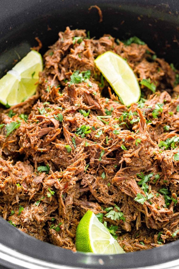 Close up photo of shredded barbacoa beef in a crockpot garnished with cilantro and lime wedges.