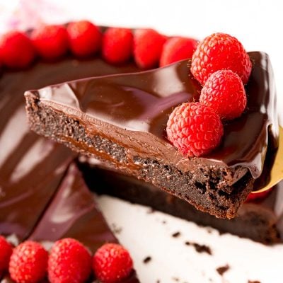Close up photo of a slice of flourless chocolate cake topped with ganache and fresh raspberries being lifted with a serving spatula from the rest of the cake.