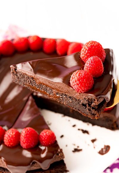 Close up photo of a slice of flourless chocolate cake topped with ganache and fresh raspberries being lifted with a serving spatula from the rest of the cake.