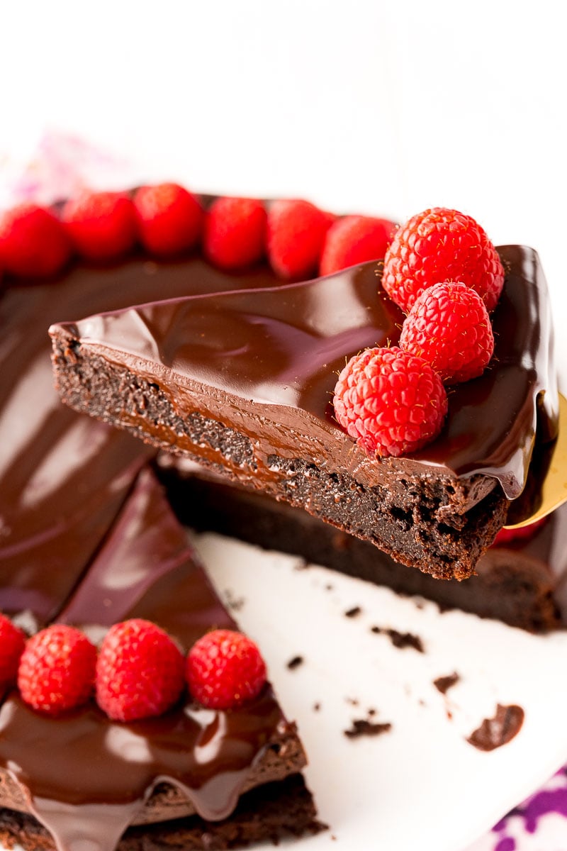 Close up photo of a slice of flourless chocolate cake topped with ganache and fresh raspberries being lifted with a serving spatula from the rest of the cake.