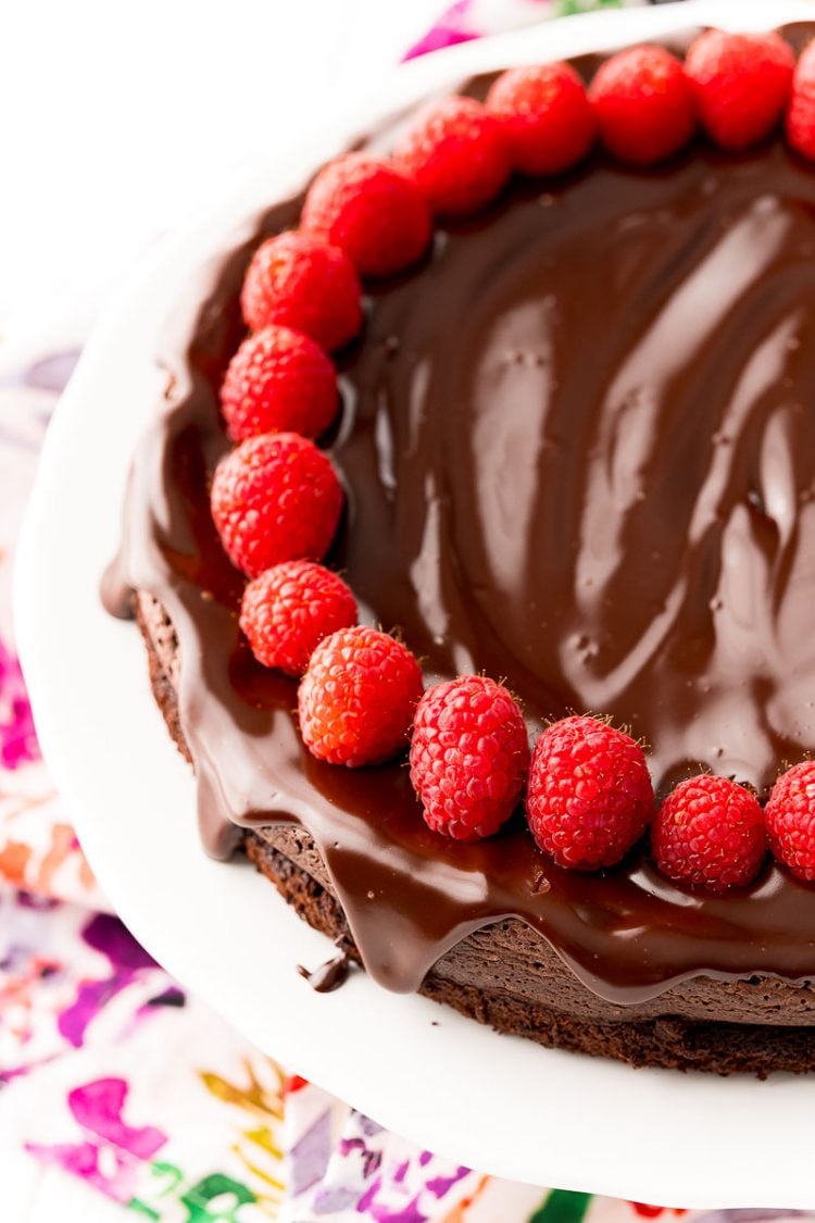 Close up photo of a chocolate cake topped with ganache and raspberries on a white cake stand.