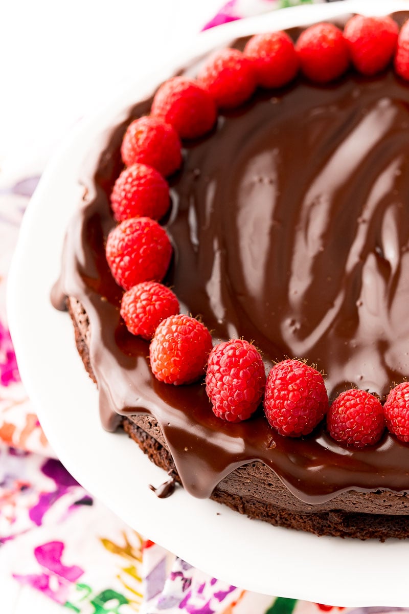 Close up photo of a chocolate cake topped with ganache and raspberries on a white cake stand.