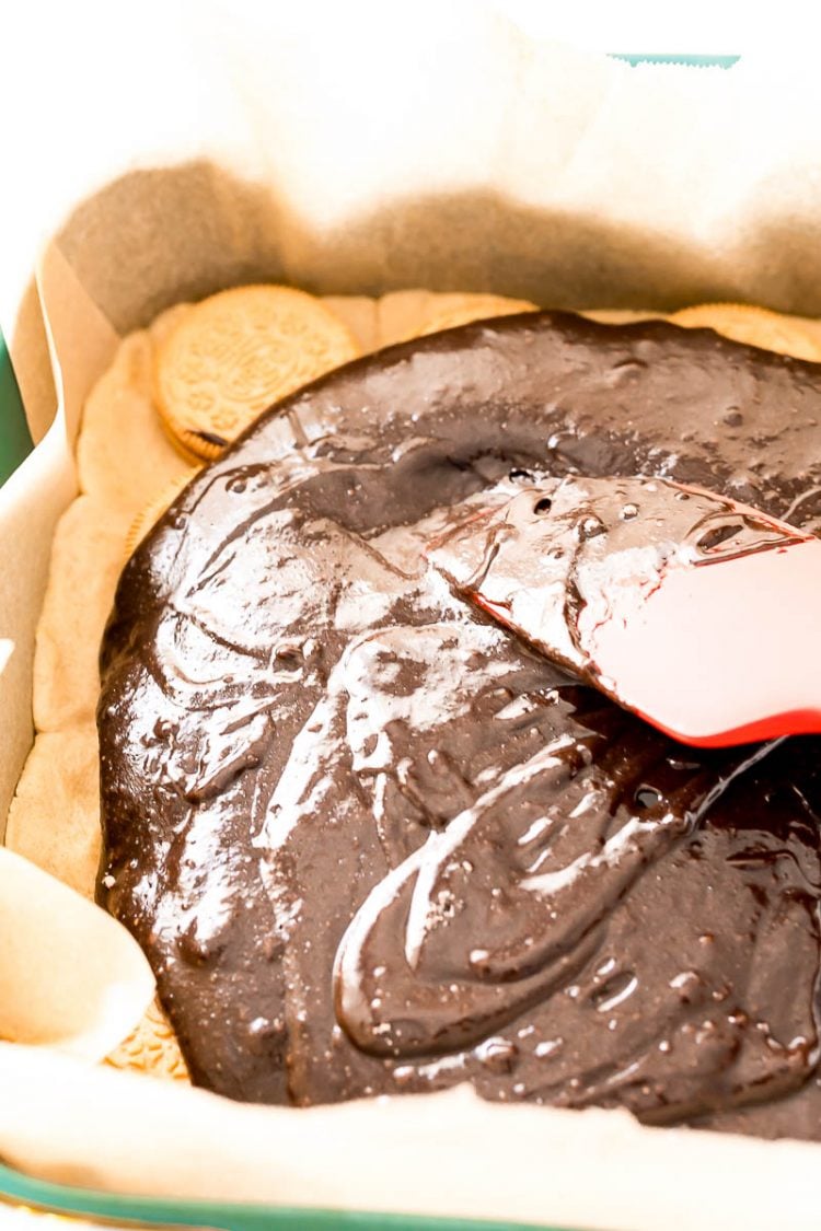 Close up photo of a rubber spatula spreading brownie batter over the top of oreos in a baking dish.
